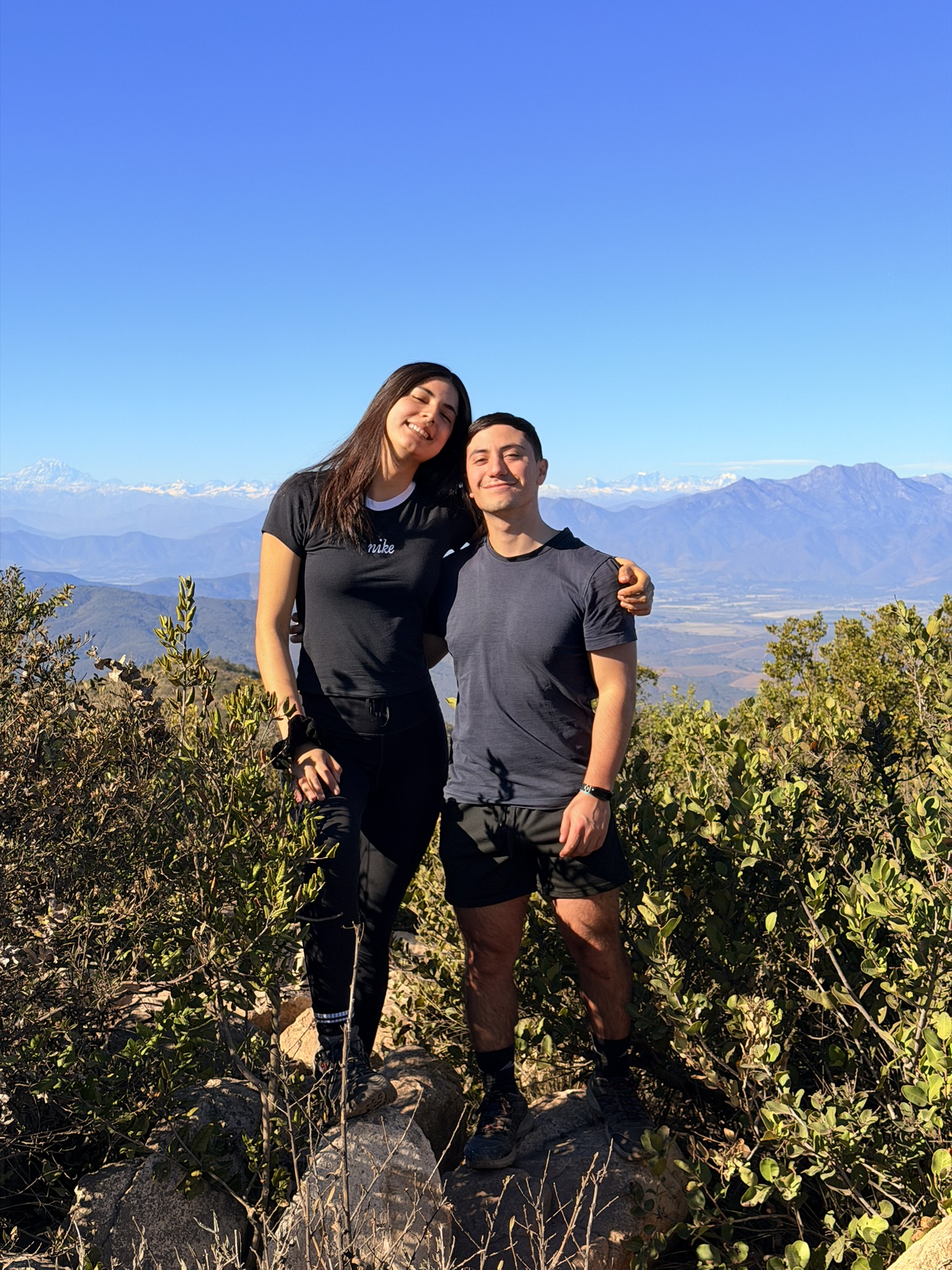 Javier y Sabrina juntos en un mirador, con montañas y cielo azul al fondo.